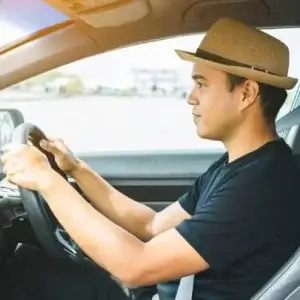 A young man with a hat driving a car with both hands on the wheel.