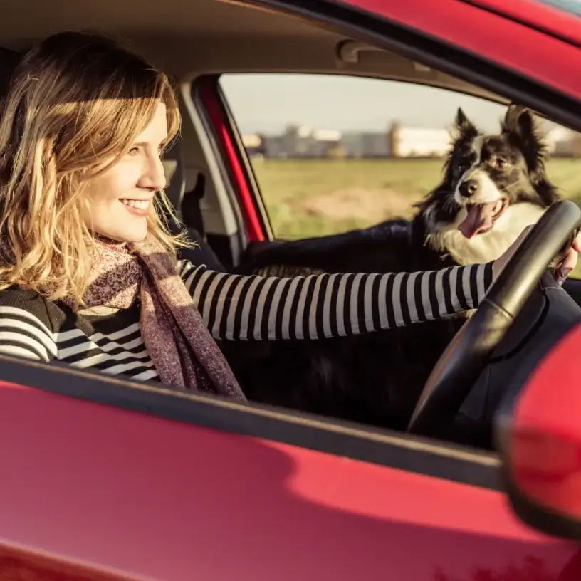 A woman driving a red car with her dog in the passenger seat.