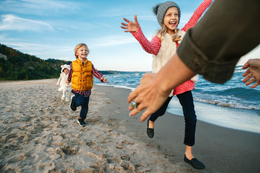 Group portrait of three funny smiling laughing white Caucasian children kids friends playing running to mother parent adult on ocean sea beach on sunset outdoors