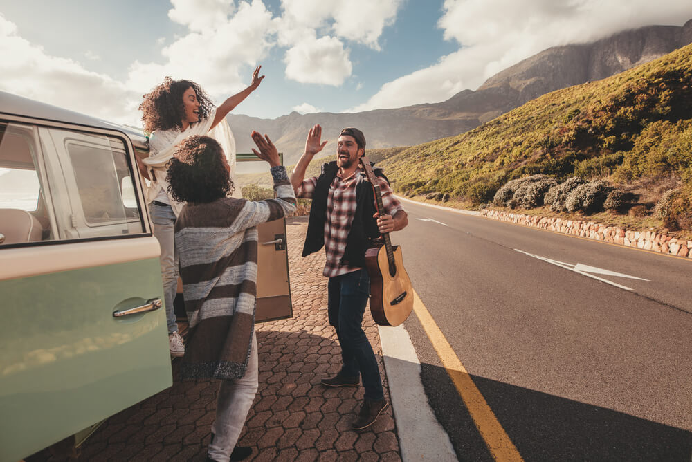 A group of friends on a road trip standing in front of a van highfiving,