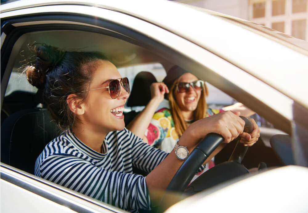 a smiling young woman driving her friend in a car on a sunny day