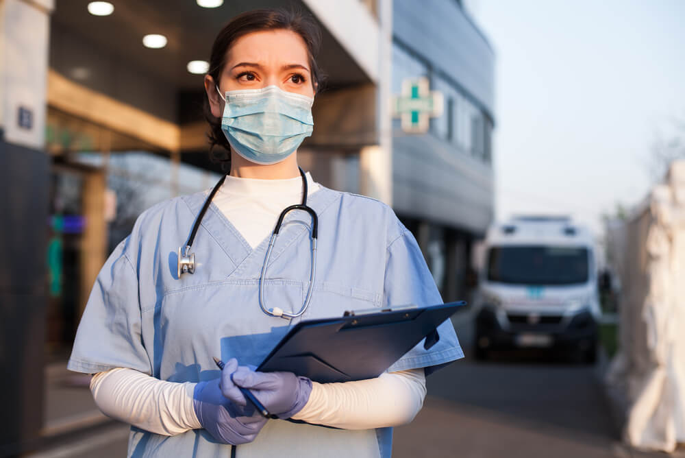 a doctor in medical scrubs outside a hospital with a clipboard and face mask