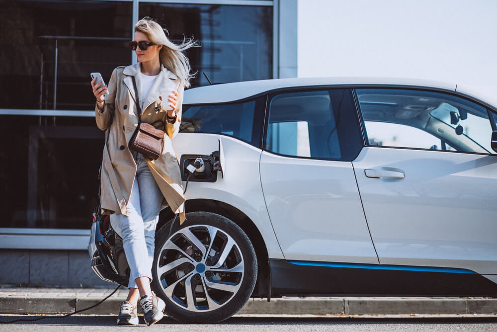 a blonde woman charging her electric car while looking at her phone