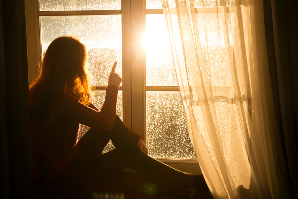 A woman sitting in windowsill looking at sun coming out after rain