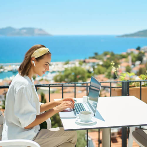 Woman on working holiday sitting with laptop on a balcony overlooking the ocean