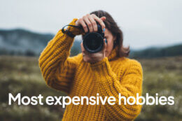 A person wearing a textured mustard‑yellow sweater holding a professional camera up to their face in an outdoor field setting, with the words ‘Most expensive hobbies’ overlaid in white text against a softly blurred landscape background.