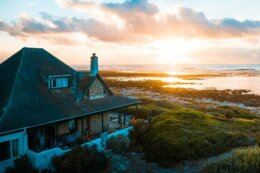 Coastal house overlooking rocky shoreline and ocean at sunset, with warm light reflecting on the water.