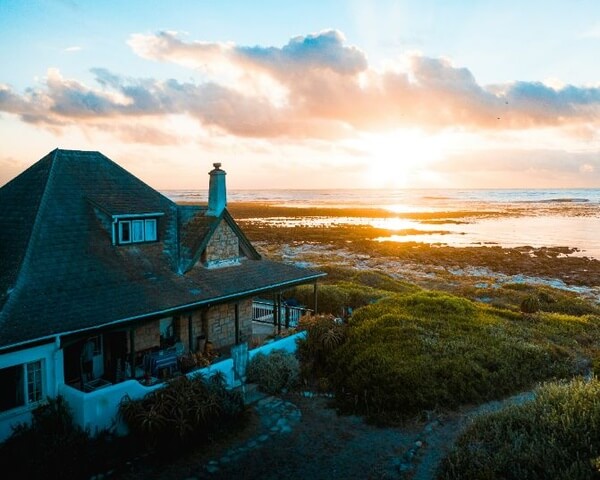 Coastal house overlooking rocky shoreline and ocean at sunset, with warm light reflecting on the water.