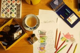 Top-down view of a wooden table with art supplies, including colored pencils, paint jars, sketchbooks with teacup illustrations, and a cup of tea.