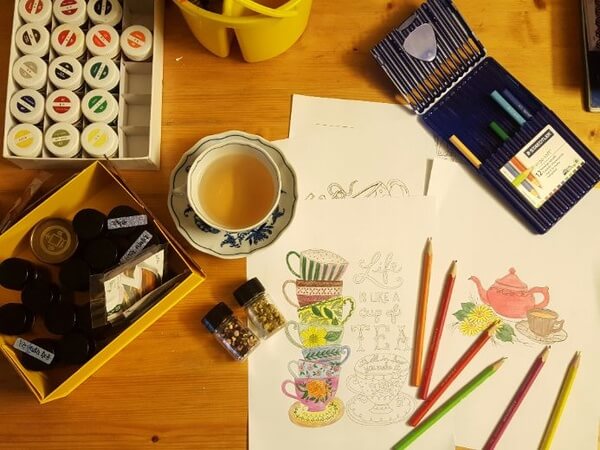 Top-down view of a wooden table with art supplies, including colored pencils, paint jars, sketchbooks with teacup illustrations, and a cup of tea.