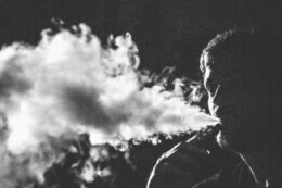 Black-and-white profile view of a person exhaling a stream of vapor/smoke into a dark background.