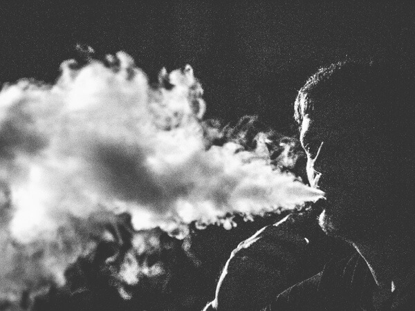 Black-and-white profile view of a person exhaling a stream of vapor/smoke into a dark background.